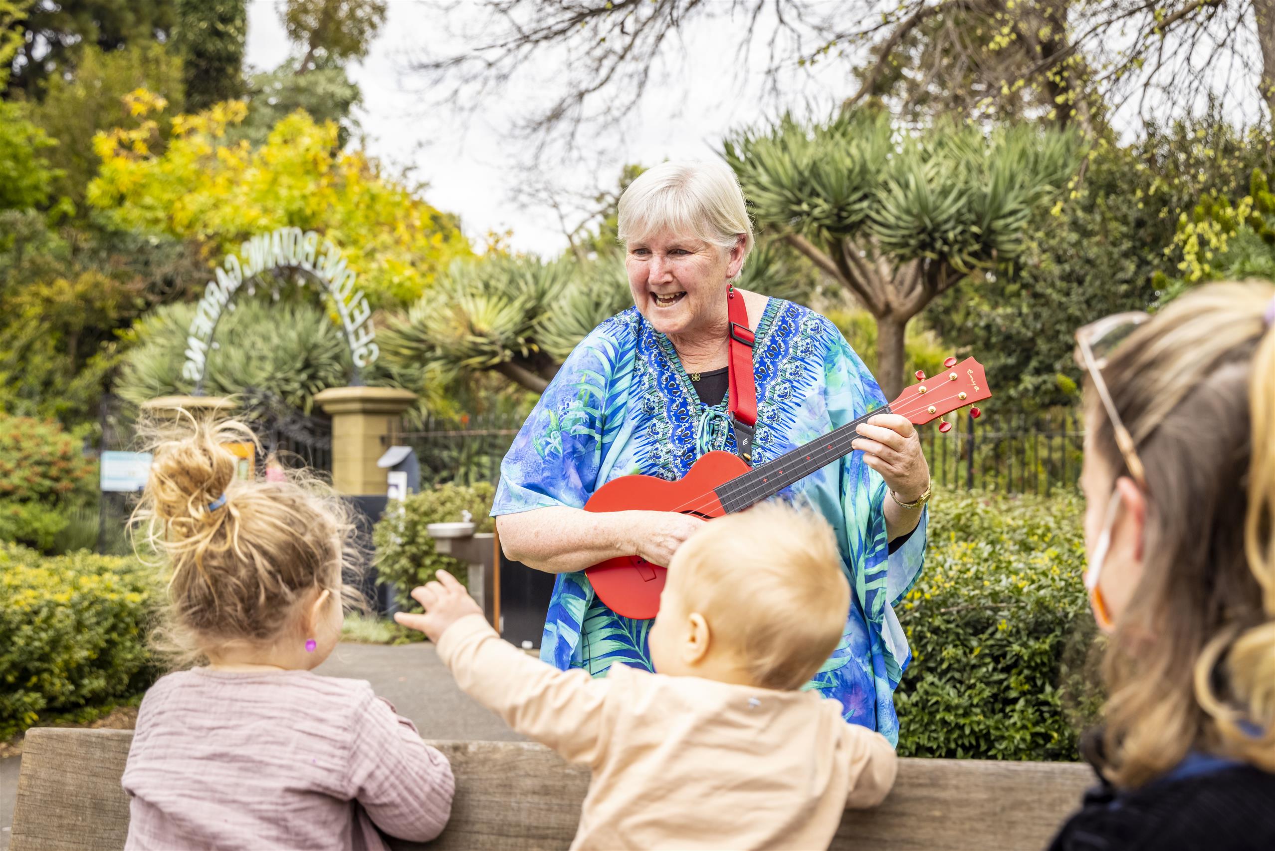 Granny Lee sings on the ukulele in the gardens to a group of young children.