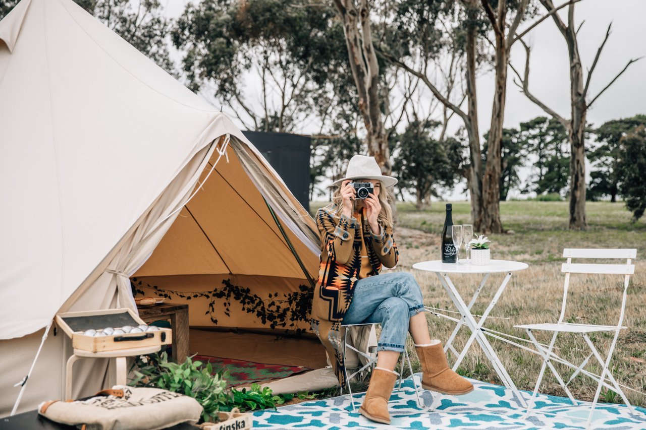 A woman sits outside a bell glamping tent with a camera in hand. Gum trees and a winery are behind the tent.