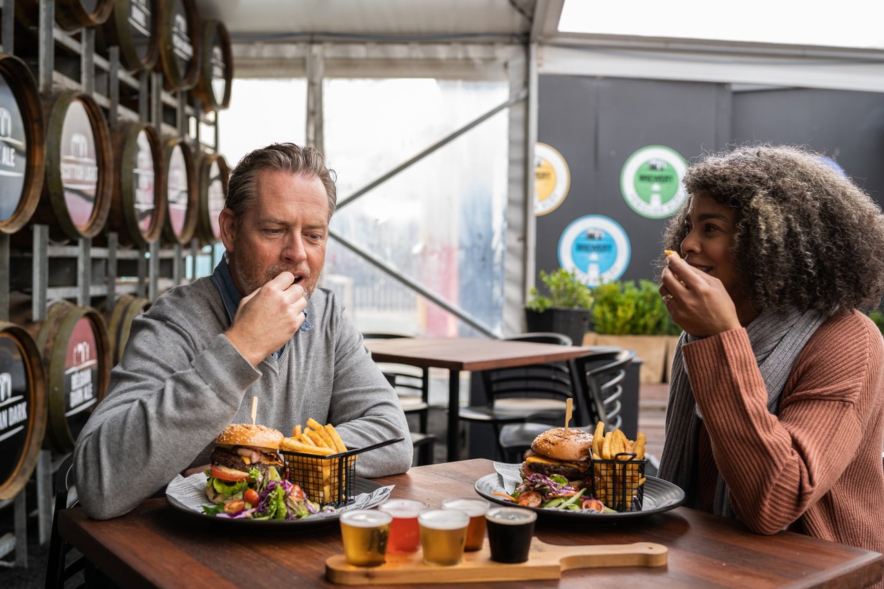 A man and woman sit at a table eating burgers with a paddle of beers from Queenscliff Brewhouse.