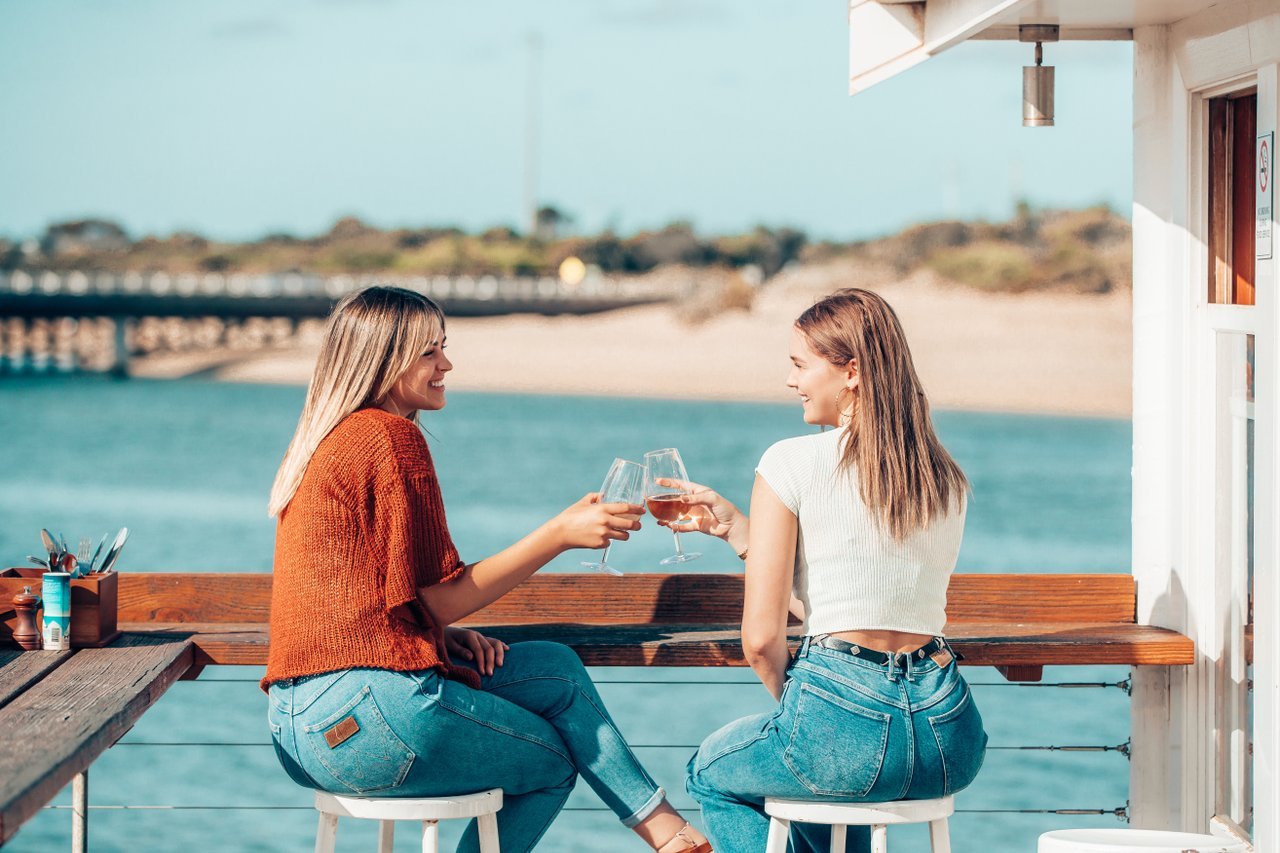 Two friends cheers glasses at coastal dining spot, At the Heads. The Barwon River is on the balcony behind them.
