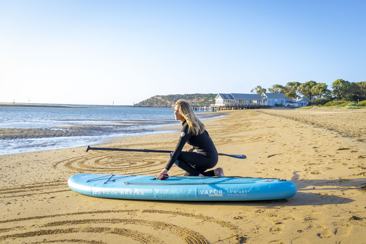 A woman crouches beside a stand up paddle board while on the sand.