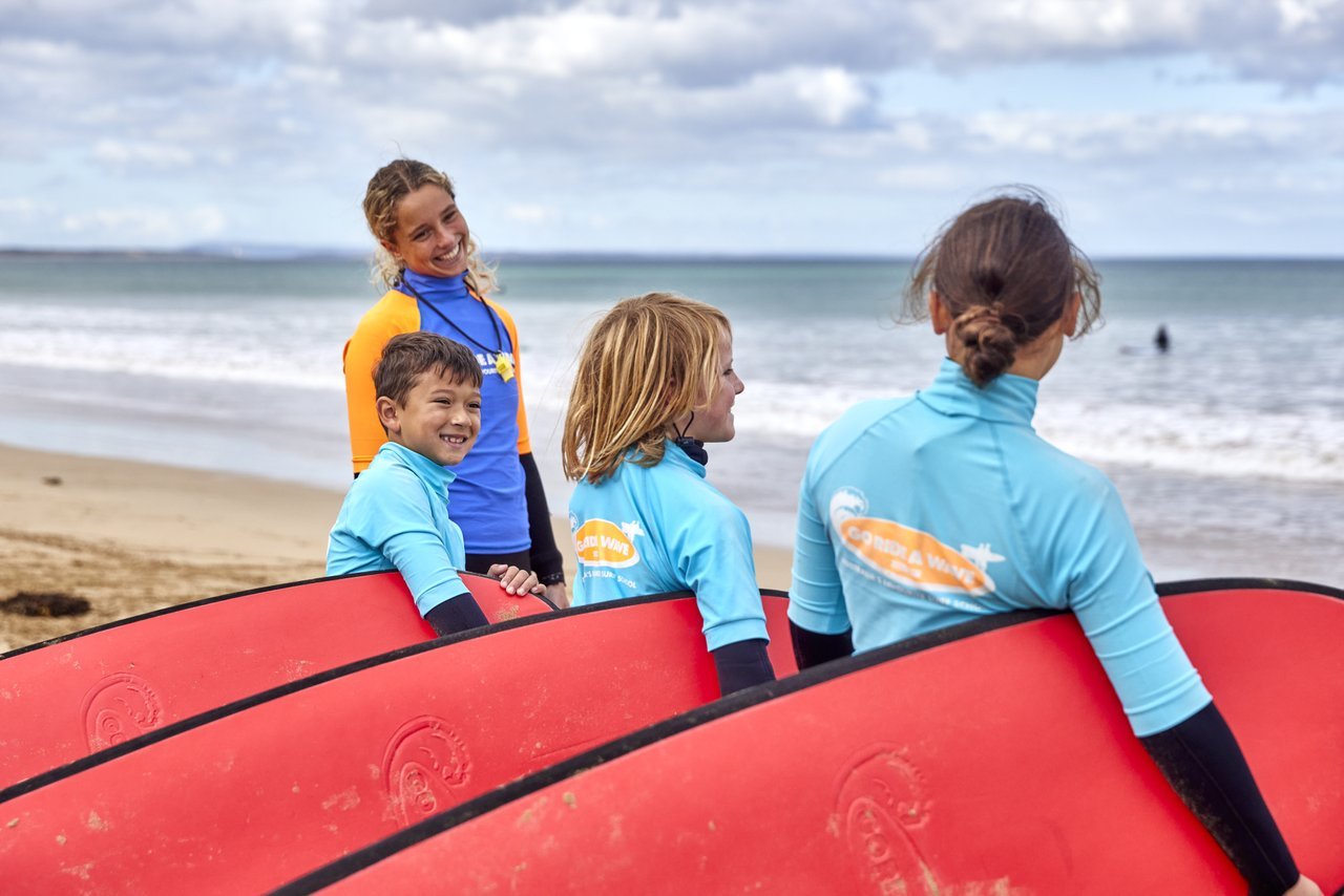 Three kids hold surf boards facing the beach, the instructor looks at them smiling.