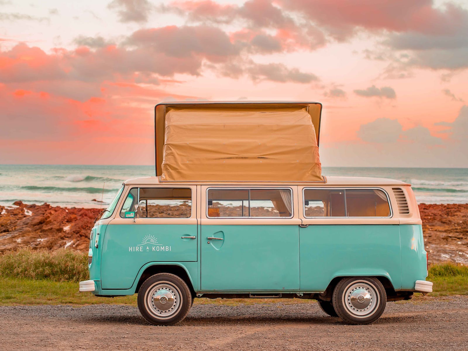 A Hire a Kombi teal coloured kombi van with its top popped beside the beach.