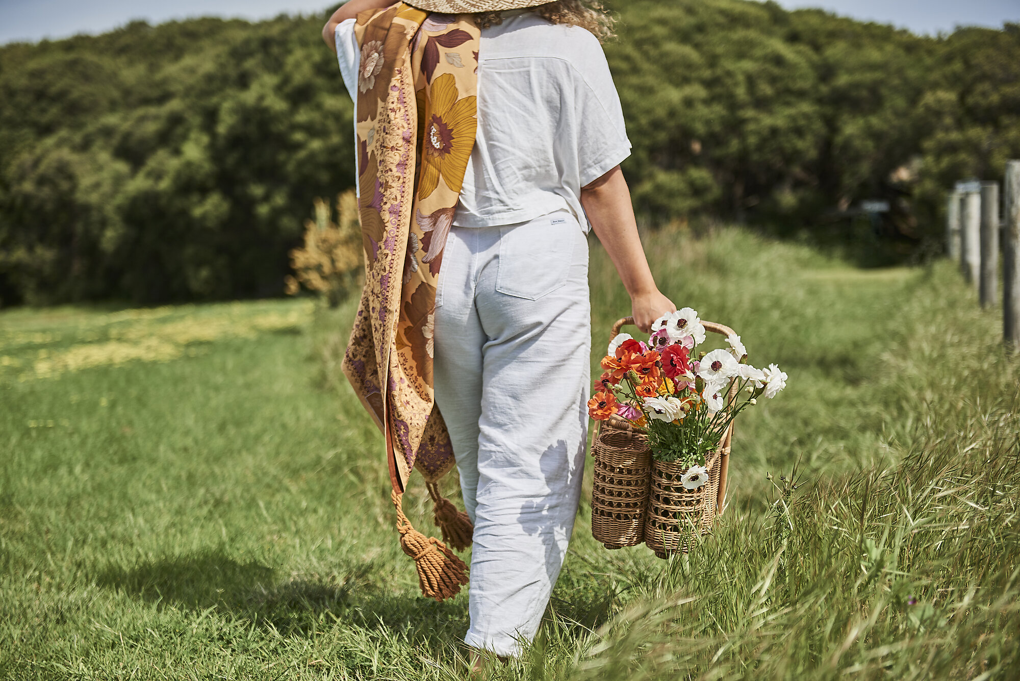 A woman walks carrying a picnic and a rug in the grass.
